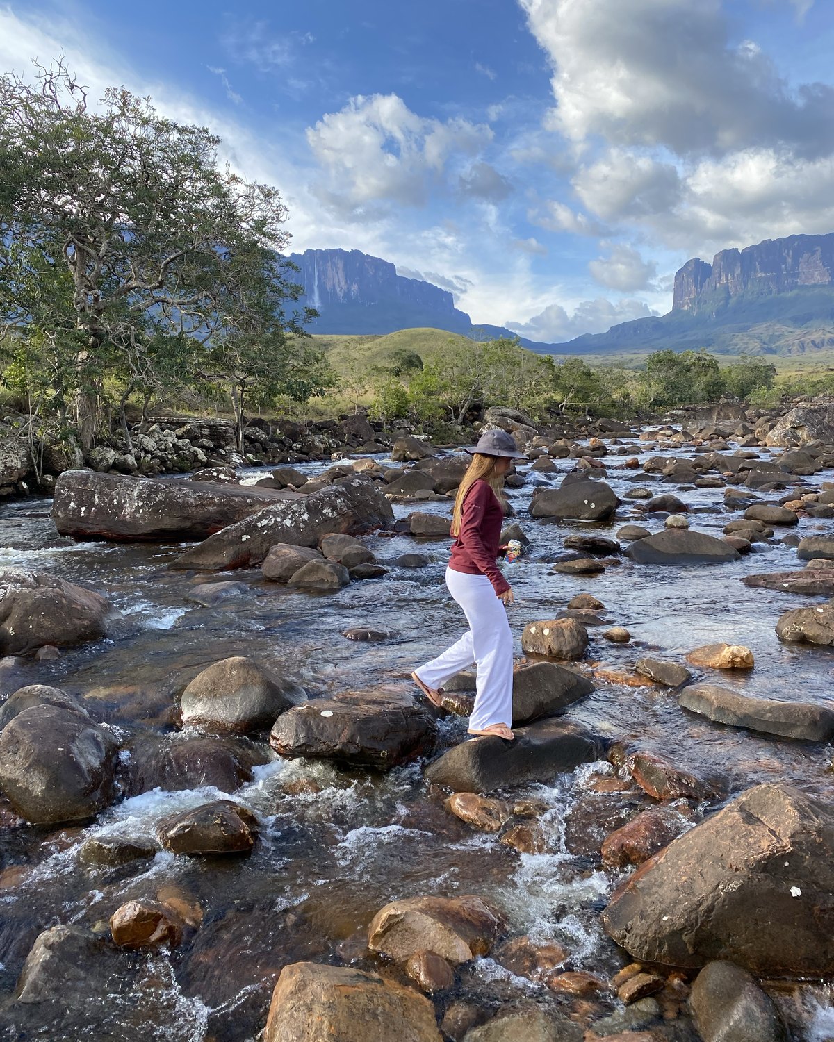 Una viajera cruzando un río de piedras con los tepuyes de la Gran Sabana al fondo